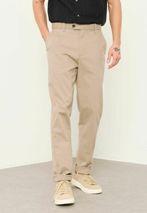 Mann trägt beige Chinos mit hochgekrempelten Säumen und beigen Turnschuhen, schwarzes kurzärmeliges Hemd teilweise sichtbar, steht mit einer Hand in der Tasche.