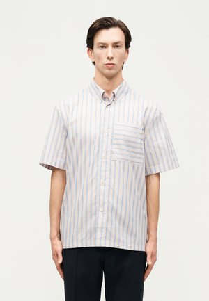 Muž v krátkym rukávom košeli s vertikálnymi pruhmi svetlomodrej a béžovej farby, stojaci pred jednoduchým svetlým pozadím.