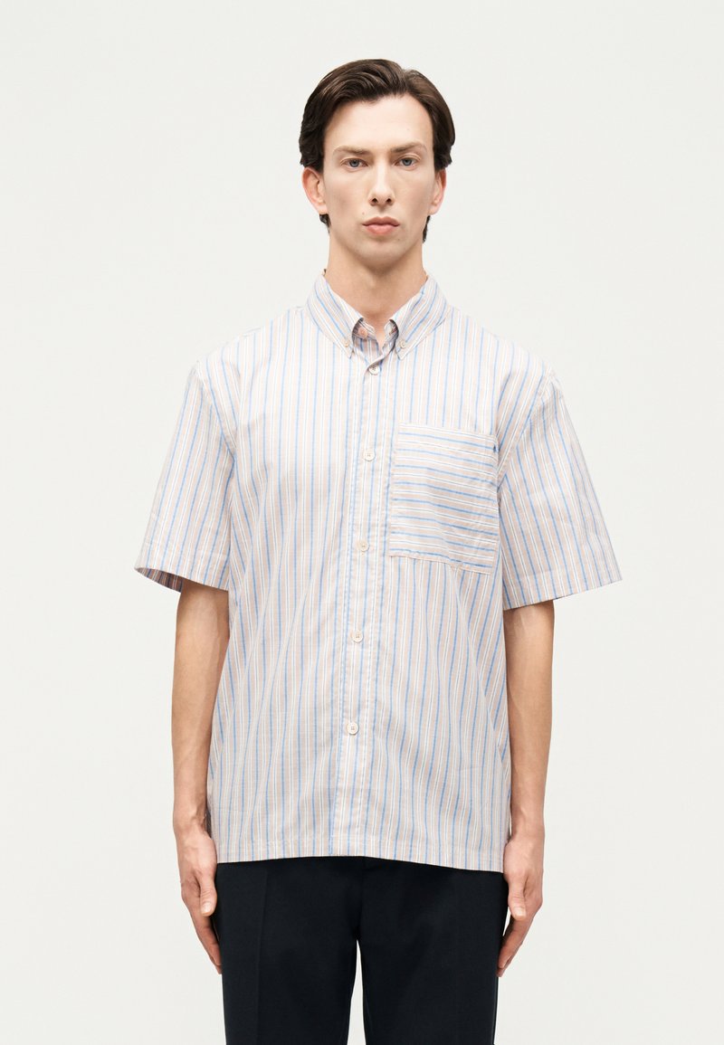 Άνδρας που φοράει πουκάμισο με κοντά μανίκια και κάθετες ρίγες σε ανοιχτό μπλε και μπεζ, στέκεται μπροστά σε απλό ανοιχτόχρωμο φόντο.