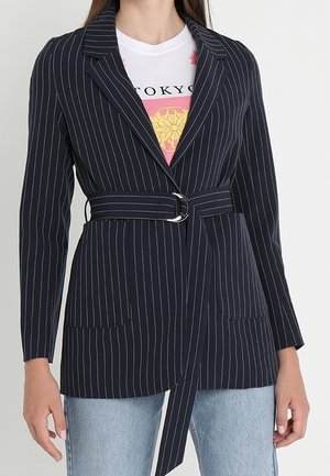 Kobieta ubrana w marynarkę w granatową prążkowaną kratę z paskiem, na której ma białą koszulkę z grafiką Tokio oraz jasnoniebieskie dżinsy.