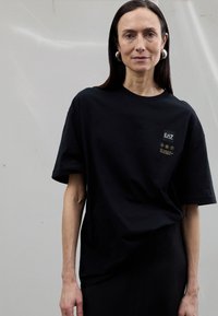 Černé oversized tričko z bavlny s krátkými rukávy, s malým logem a grafikou na hrudi. Hladká textura.