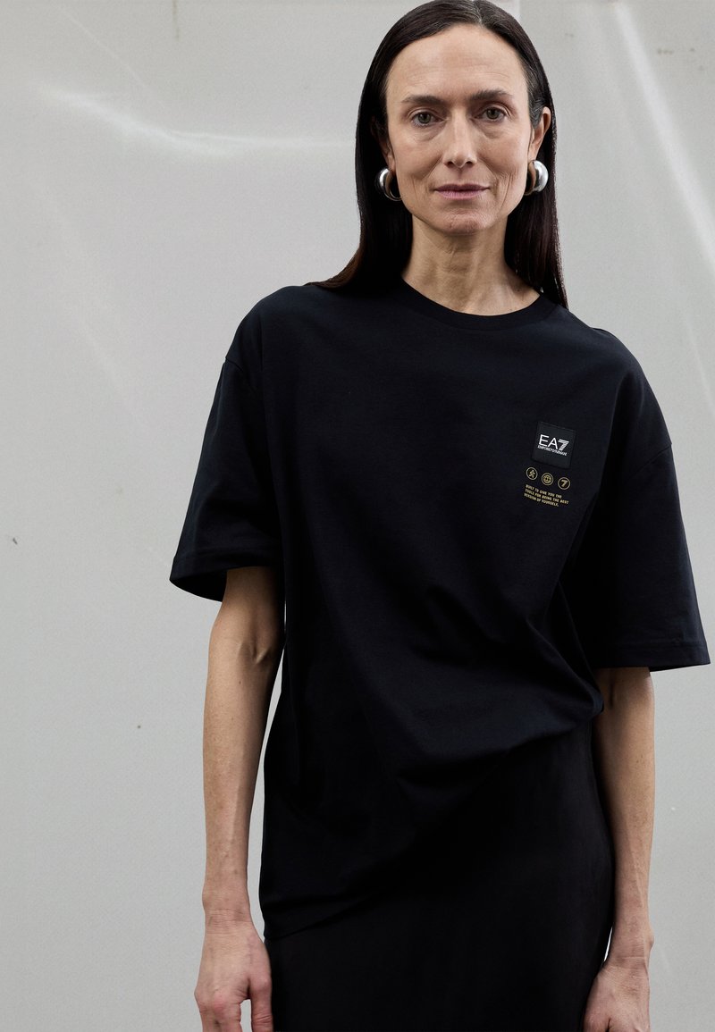 Černé oversized tričko z bavlny s krátkými rukávy, s malým logem a grafikou na hrudi. Hladká textura.