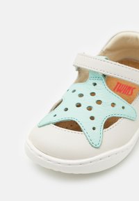 Valkoinen nahkakenkä, jossa on turkoosi tähden leikkauskuvio, varustettuna rei'ityksillä ja Velcro-hihnalla. Pehmeä sisäpinta mukavuutta varten.
