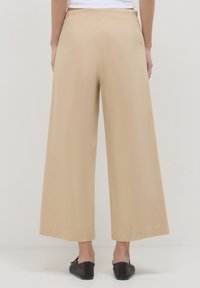 Persona che indossa pantaloni beige a gamba larga e scarpe flats nere, vista da dietro su uno sfondo bianco uniforme.
