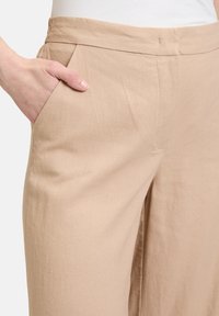 Beige weit geschnittene Hose aus weichem Stoff, mit glatter Textur, seitlicher Tasche und tailliertem Bund mit minimalen Nähten.