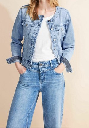 Vrouw draagt een lichtblauwe spijkerjas over een wit hemd, gecombineerd met hooggetailleerde losse blauwe jeans, handen in de zakken.