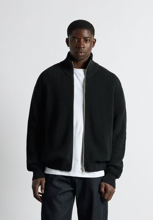 Jeune homme portant un pull noir côtelé avec fermeture éclair sur un t-shirt blanc et un pantalon foncé, debout devant un fond clair uni.