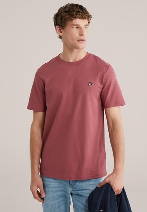 Jonge man die een roestachtige roze t-shirt en lichtblauwe spijkerbroek draagt, met een marineblauwe jas in zijn linkerhand, terwijl hij naar rechts kijkt.