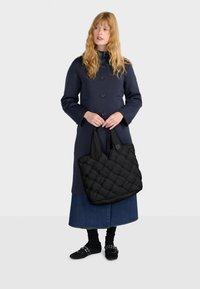 Borsa a spalla nera trapuntata con texture imbottita e manici larghi, abbinata a un cappotto blu navy, una gonna di denim e scarpe nere.