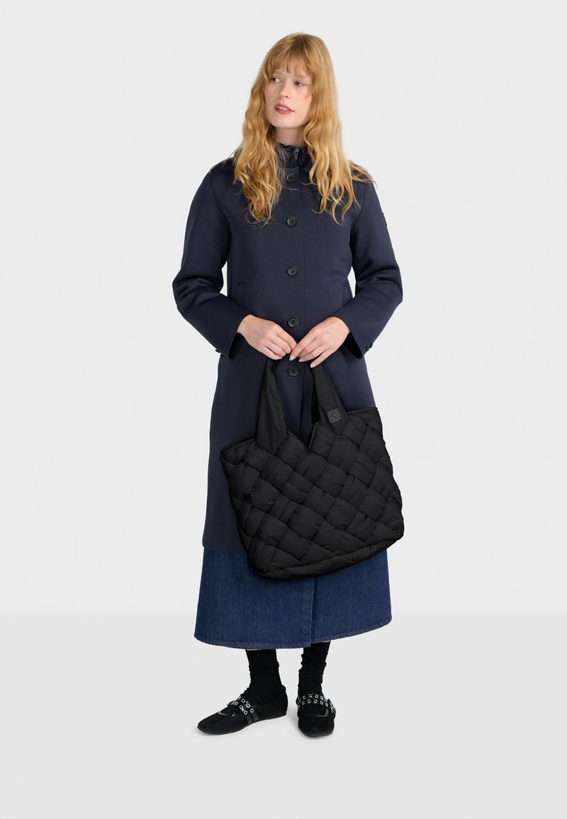 Borsa a spalla nera trapuntata con texture imbottita e manici larghi, abbinata a un cappotto blu navy, una gonna di denim e scarpe nere.