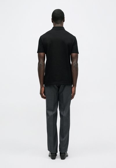 Juoda polo marškinėliai trumpomis rankovėmis, derinami su pilkais, tekstūruotais oficialiais kelnėmis, dėvimi asmens, matomo iš nugaros.