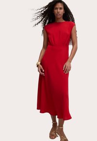 Abito rosso fino al ginocchio con corpetto aderente e leggero volant sull'orlo, realizzato in tessuto liscio. La modella indossa accessori dorati e sandali.