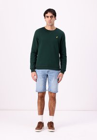 Felpa verde scuro con polsini e orlo a costine, abbinata a pantaloni shorts in denim azzurri. L'outfit include sneakers marroni e calzini bianchi.