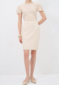 Top beige con textura de canalé y mangas abullonadas combinado con una falda lápiz. Textura suave, hasta la rodilla. Lleva unos tacones beige de punta afilada y una pulsera.