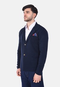 Cardigan lavorato a maglia blu navy con due tasche frontali, colletto a revers e bottoni marroni. Presenta un fazzoletto da taschino a fantasia nella tasca sinistra del petto.