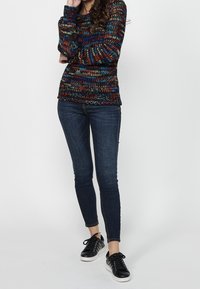 Meerbruine gebreide trui met een gestructureerd patroon, gecombineerd met donkere skinny jeans en zwarte sneakers met witte zolen en zilveren accenten.