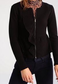 Kvinna som bär en svart texturerad zip-up jacka med volangdetaljer över en brun leopardmönstrad genomskinlig topp och mörka jeans.