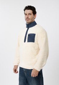 Φλις επάνω μέρος σε κρεμ απόχρωση με ψηλό ναυτικό γιακά, τσέπη στο στήθος και κλείσιμο με φερμουάρ στη μέση. Διαθέτει μαλακή, υφή επιφάνεια.