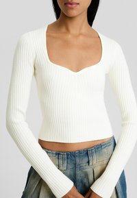 Crop top blanc à manches longues côtelées avec un décolleté carré, une coupe ajustée et une texture subtile. Associé à un pantalon en denim bleu clair taille haute.