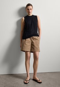 Schwarzes ärmelloses Top kombiniert mit khakifarbenen, gefalteten Shorts, aus strukturiertem Stoff, mit schwarzen Flip-Flops, vor einer hellgrauen Wand.