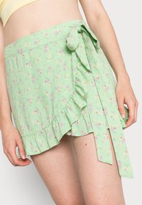 Saia envolvente floral verde com uma bainha franzida, apresentando pequenas flores rosas e amarelas. Inclui um detalhe de laço lateral e é feita de um tecido leve e macio.