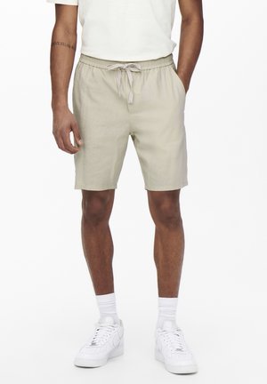Shorts beiges à cordon de serrage en tissu léger, dotés de poches latérales et d'une coupe décontractée, portés avec un t-shirt blanc et des baskets.