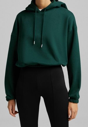 Personne portant un sweat à capuche vert foncé avec des cordons et des manches longues, associé à un legging noir à côtes taille haute.