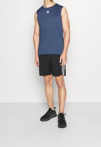 Tmavě modrý sportovní top bez rukávů s kulatým výstřihem a logem Adidas, doplněný černými šortkami s bílými pruhy, nošený s černými teniskami.