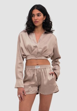 Kvinde med krøllet mørkt hår iført en beige satin kort knap-op skjorte og matchende elastiske taljeshorts med etiketten "WRSTBHVR".