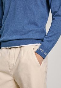 Blå stickad tröja med ribbad nederkant och mudd, med lätt broderi på muddarna. I kombination med beige byxor, ficka synlig.