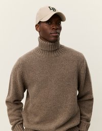 Καφέ πλεκτό πουλόβερ με ψηλό γιακά, συνδυασμένο με μπεζ καπέλο που φέρει σκούρο πράσινο κείμενο, σχεδιασμένο με χαλαρή εφαρμογή.