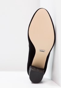 Chaussure à talon haut en suède noir avec un talon texturé et pailleté et une semelle en caoutchouc lisse. Elle présente un bout pointu et des détails de couture subtils.