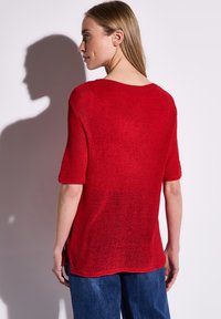 Rotes, kurzärmeliges Strickoberteil mit lockerer Passform, Rundhalsausschnitt und seitlichen Schlitzen. Die Textur ist weich und leicht durchsichtig, mit einer glatten Strickoberfläche.