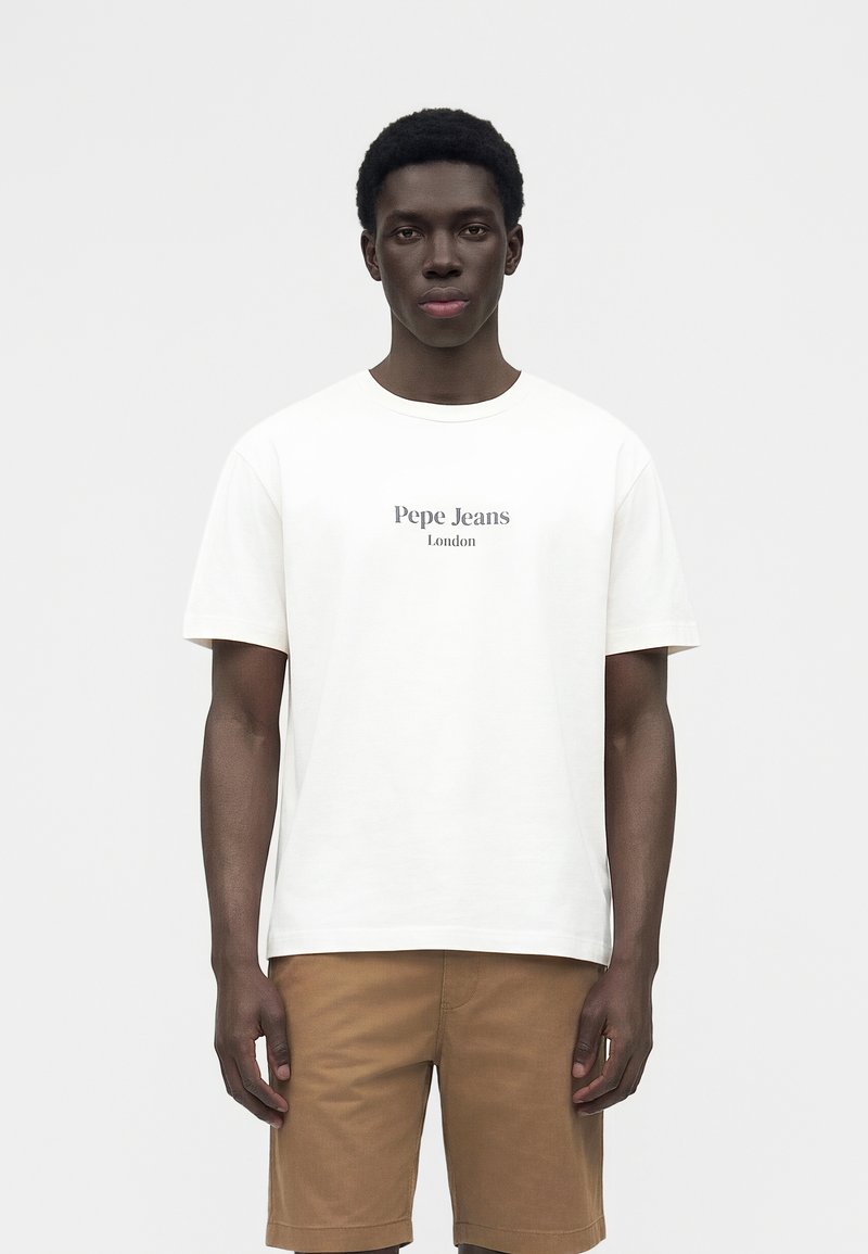 Νέος Μαύρος άντρας που φοράει λευκό T-shirt Pepe Jeans London και καφέ σορτς, στέκεται μπροστά σε μια απλή φωτεινή επιφάνεια.