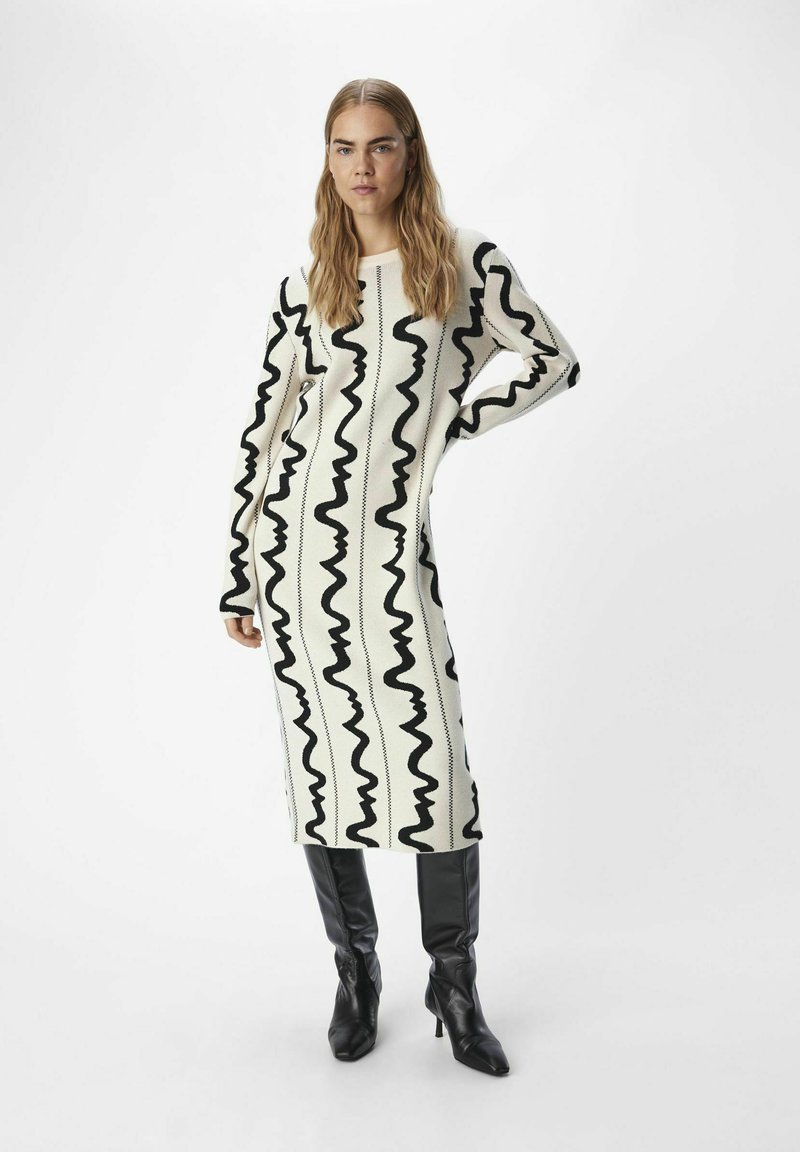 Langes Strickkleid in Creme mit schwarzem Wellenmuster, rundem Ausschnitt und langen Ärmeln. Kombiniert mit kniehohen schwarzen Stiefeln mit Absatz.