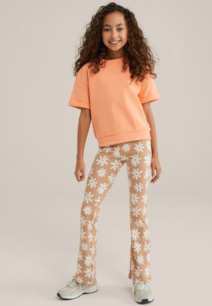 Oranje sweatshirt met korte mouwen, gecombineerd met beige wijde broek met witte bloemen en een lichtroze midden, aangevuld met grijze sneakers.