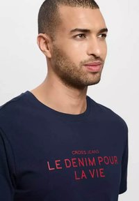 Granatowa koszulka z okrągłym dekoltem i krótkimi rękawami; z przodu z wyraźnym czerwonym napisem: "CROSS JEANS LE DENIM POUR LA VIE".