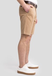 Khaki-Shorts mit geradem Schnitt, seitlichen Taschen und genähtem Saum. Getragen mit weißen Sneakers mit brauner Sohle und Schnürdesign.