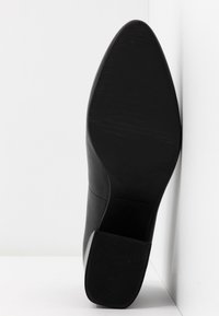 Semelle de chaussure noire à bout pointu avec un talon carré appuyée contre un coin blanc entre un mur et le sol.