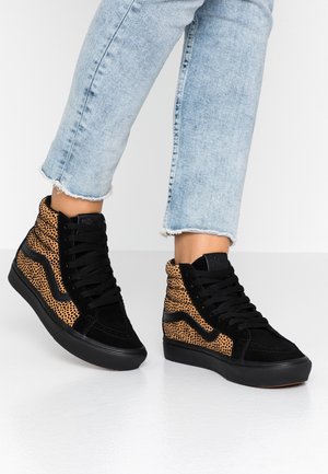 Osoba ubrana w jasnoniebieskie skrócone dżinsy i czarne wysokie trampki z brązowymi cętkowanymi panelami, stojąca na białej podłodze.