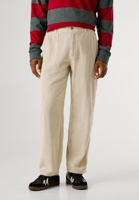 Bežīgas korduroja bikses ar vertikālām svītrām, kam ir poga jostasvietā. Apvienotas ar melniem sporta apaviem un pelēku un sarkanu svītrotu krekliņu.