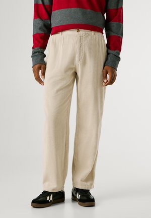 Pantaloni in corduroy beige con righe verticali, dotati di un bottone in vita. Abbinati a sneaker nere e a una maglietta a righe grigia e rossa.