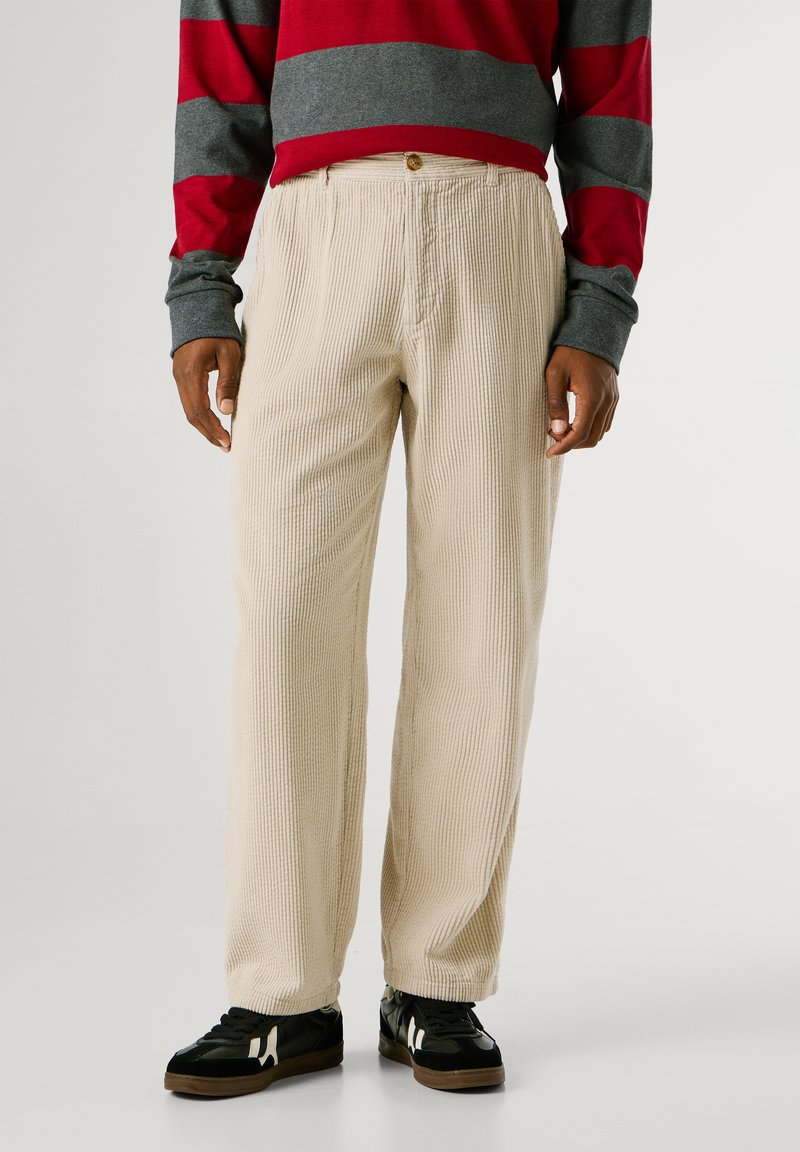 Bežīgas korduroja bikses ar vertikālām svītrām, kam ir poga jostasvietā. Apvienotas ar melniem sporta apaviem un pelēku un sarkanu svītrotu krekliņu.