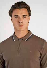 Brun zip-polo tröja med en texturerad tyg, som har en kontrasterande krage med blå och vita ränder samt en liten logotyp på bröstet.