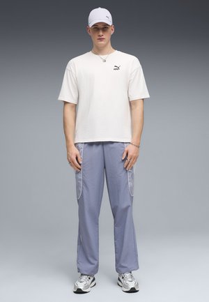 Joven de pie con gorra blanca de Puma, camiseta blanca, pantalones cargo azul claro con bolsillos de malla y zapatillas plateadas, sobre un fondo degradado gris.