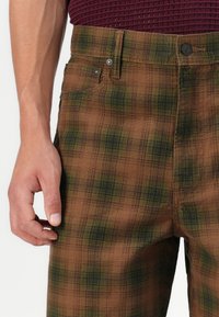 Brune ternede bukser med et diskret grønt mønster, standard fem-pockets design og metalhardware i taljen samt foran knapperne.
