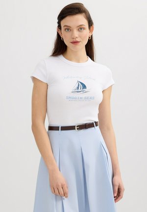 Joven mujer de cabello largo y castaño lleva una camiseta blanca ajustada con gráfico de velero azul y falda plisada azul clara con cinturón marrón.