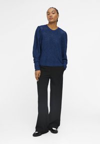 Top in pizzo lavorato a maglia, di colore blu navy e maniche lunghe, abbinato a pantaloni neri a gamba larga e scarpe nere con dettagli bianchi.