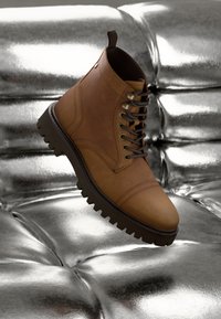 Bota de cuero marrón con cordones y suela gruesa flotando sobre una superficie metálica plateada brillante y acolchada.