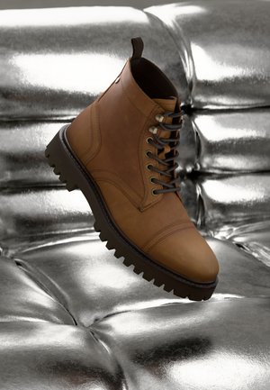 Bota de cuero marrón con cordones y suela gruesa flotando sobre una superficie metálica plateada brillante y acolchada.